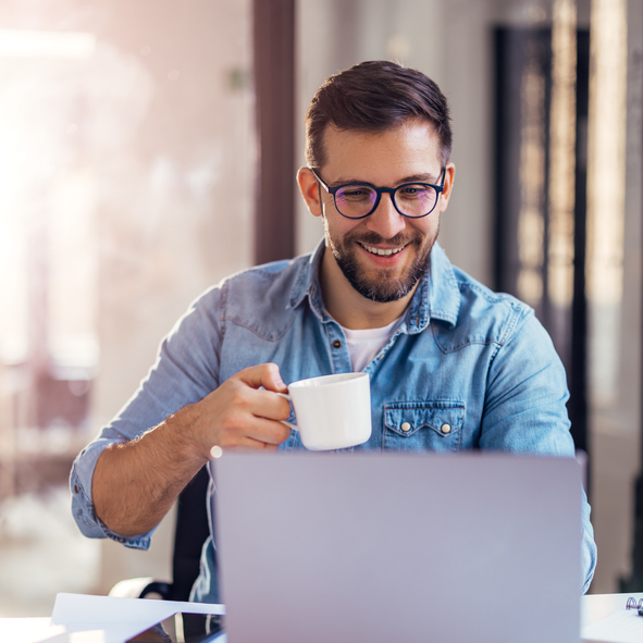 Handsome smiling young man sitting by window in office, drinking coffee while working on laptop. Job Alerts
